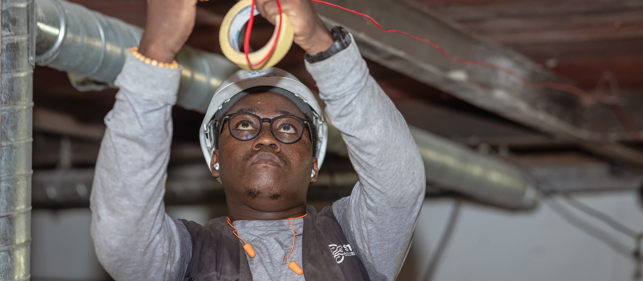 An electrician in a hard hat running red cable along a ceiling joist on site