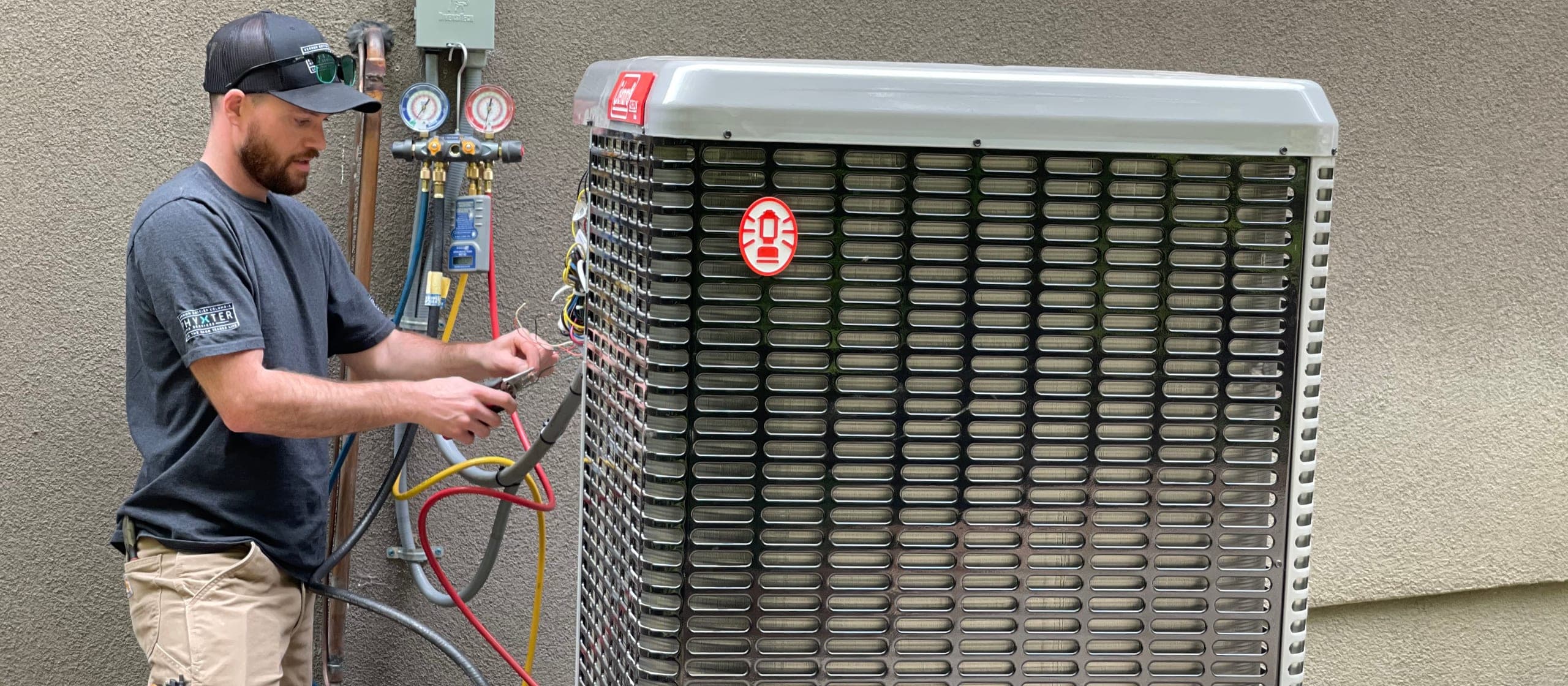 An HVAC technician with gauges on the manifold, wiring an outdoor condenser unit beside a house