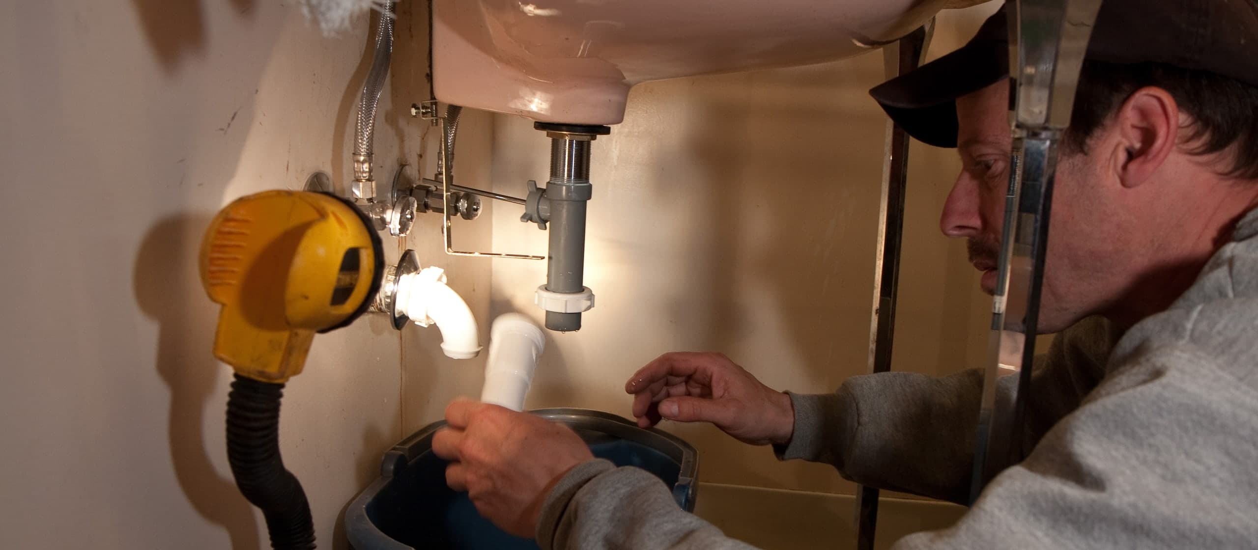 A plumber in a cap working under a bathroom sink, draining the trap into a bucket