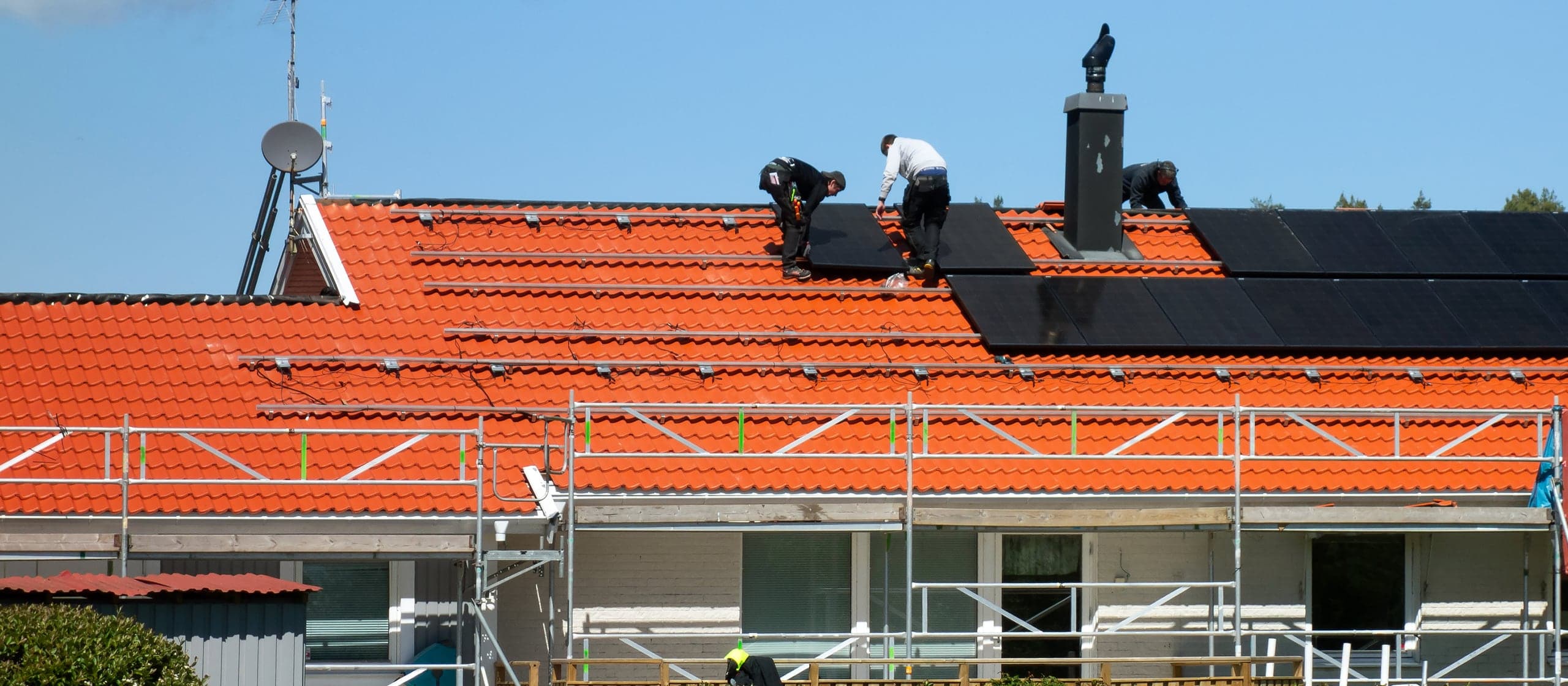 Three installers laying solar panels across the tiled roof of a house, with scaffolding along the eaves